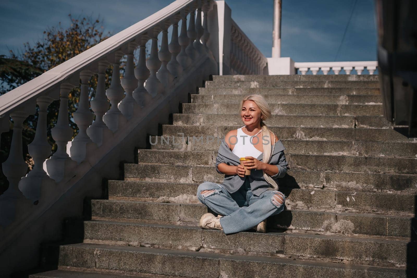 A woman sits on a set of stairs, holding a cup of coffee. She is enjoying her coffee and taking in the view from the top of the stairs