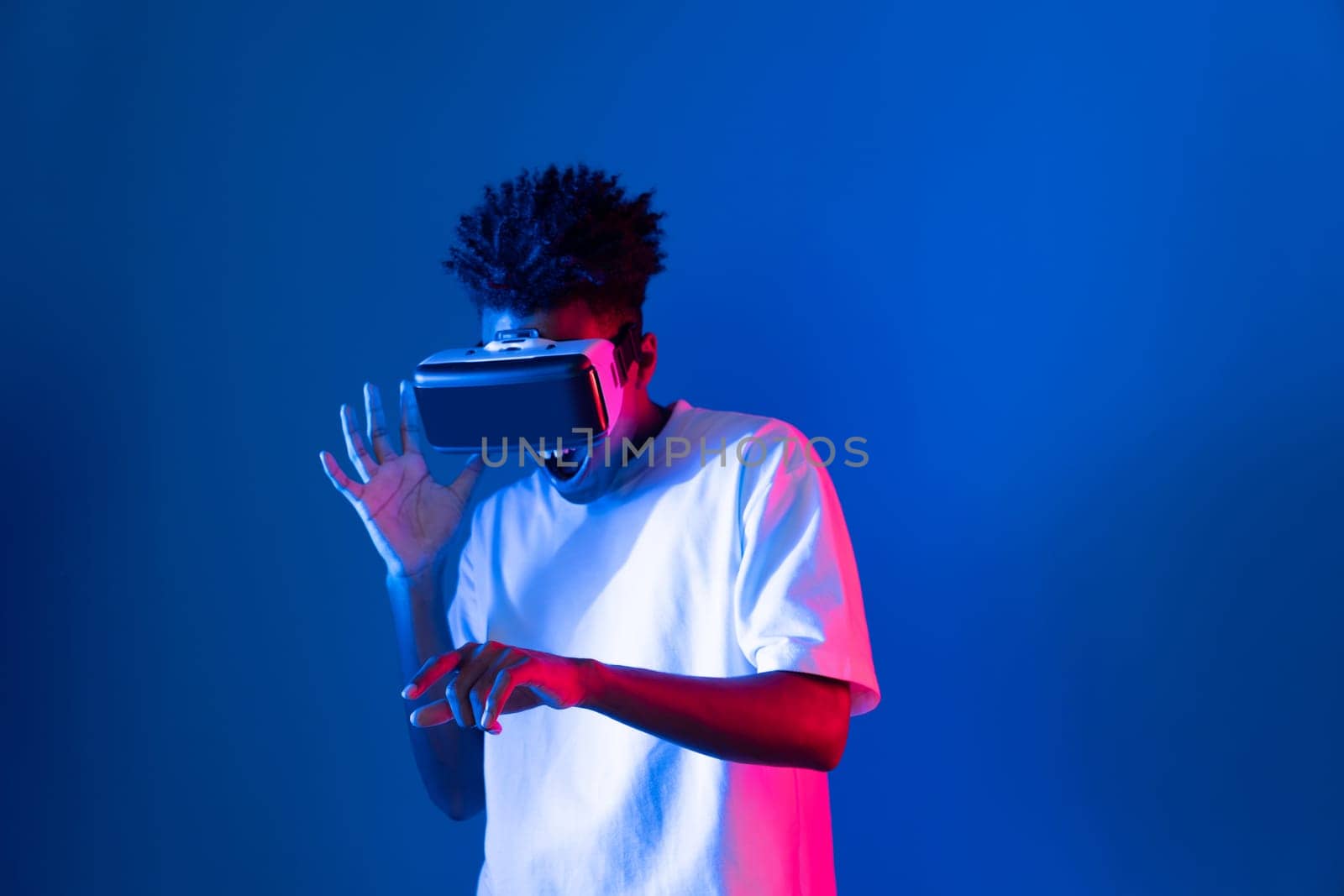 Young African American gaming player wearing VR pointing hologram 3D graphic isolated blue pink neon light wall connecting digital futuristic technology virtual reality metaverse world. Contrivance.