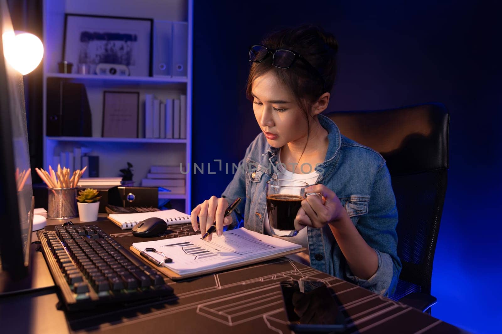 Thinking young beautiful Asian creative woman looking on pc with holding coffee cup, pointing sales target report on business market graph folder paperwork report at neon at night time. Stratagem.