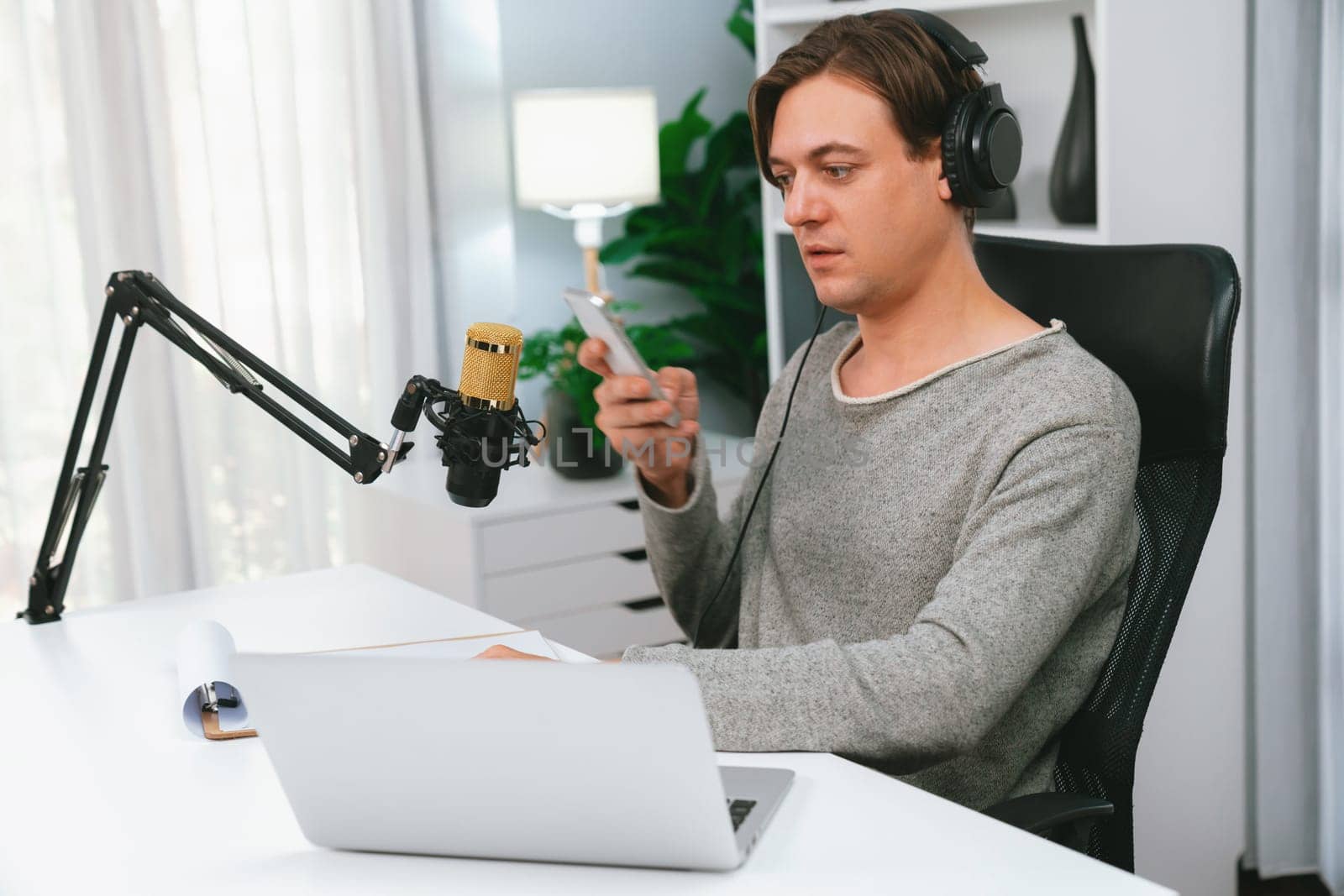 Host channel in smiling smart broadcaster recording, talking show with script and smartphone on desk on live social media, wearing headphones to record video streamer at modern home studio. Pecuniary.