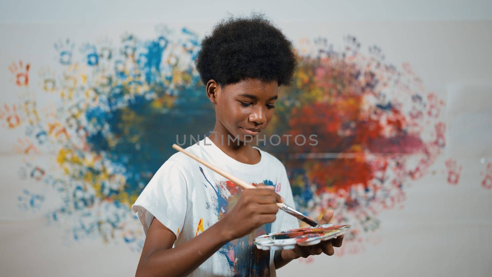 African boy standing at colorful stained wall while holding brush and point at camera. Happy highschool student wearing white shirt with stain color and look at camera with confident. Edification.