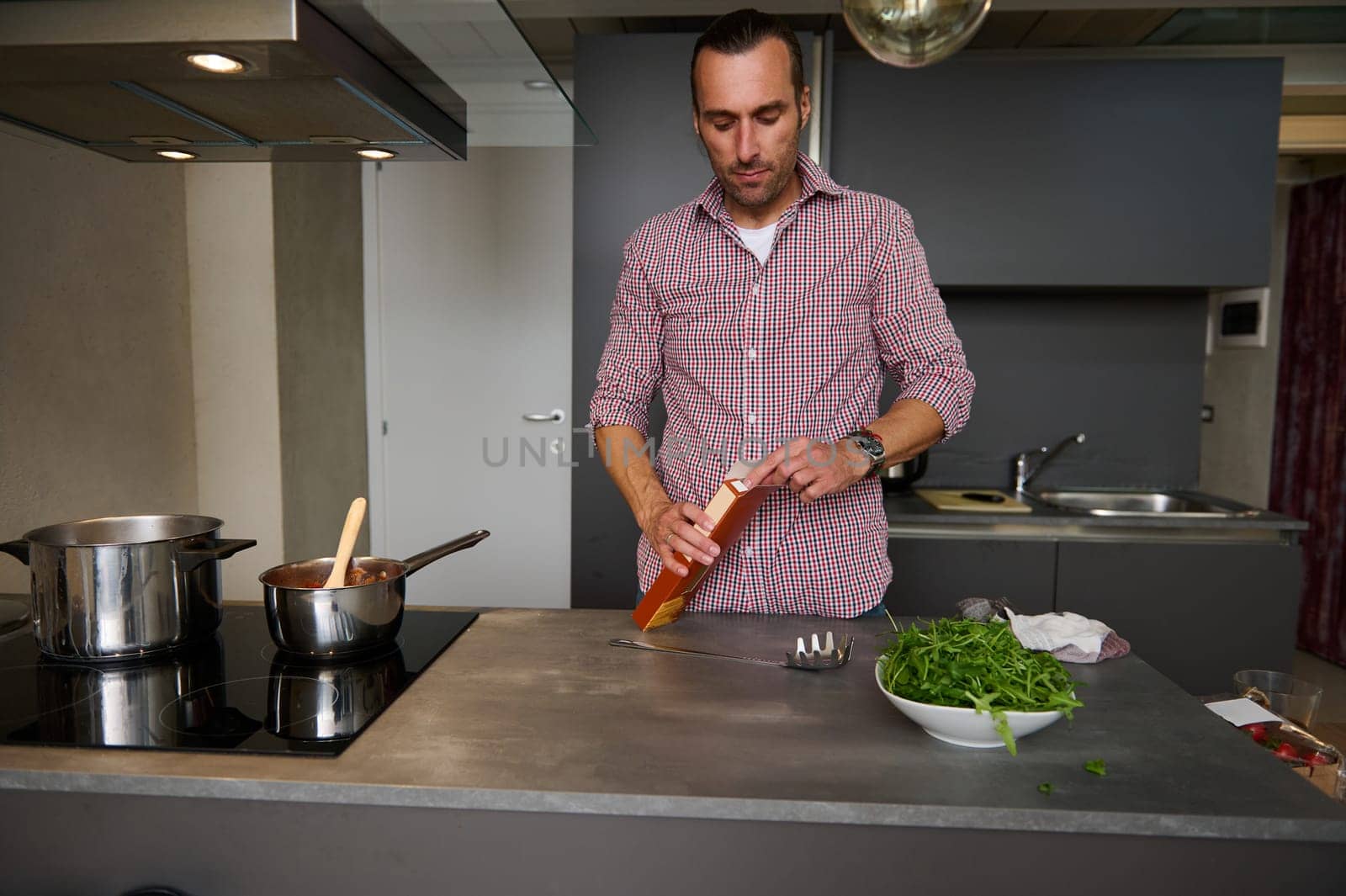 Young man chef opening a cardboard pack with integral Italian pasta, standing at kitchen counter in modern minimalist home kitchen interior. People. Lifestyle. Italian food and cuisine