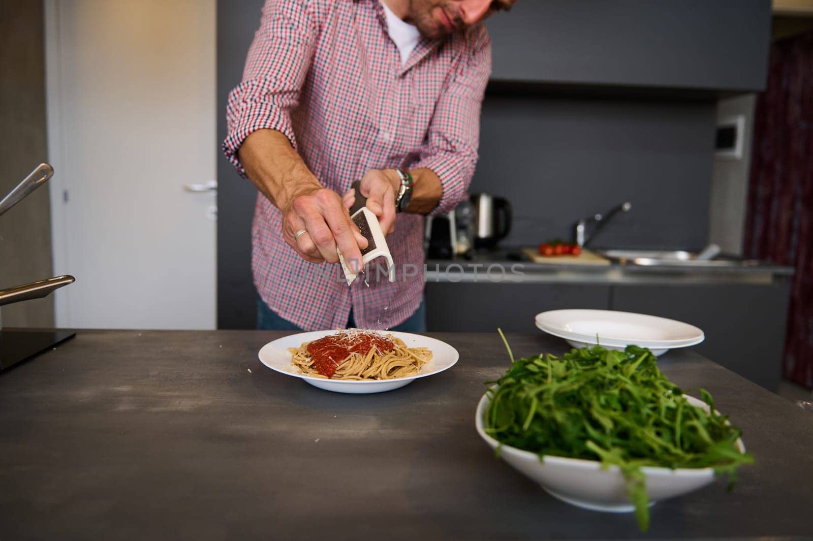 European man cooking pasta for dinner at home. Close-up hands holding a grater, grating cheese while seasoning the freshly boiled spaghetti, preparing healthy meal according to Italian recipe