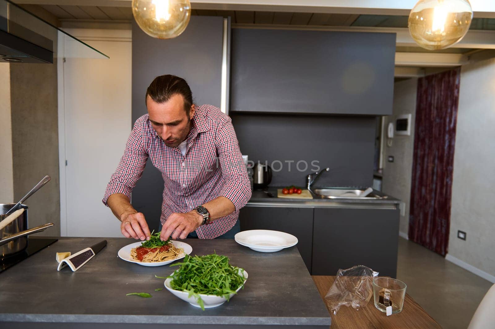 Young Caucasian man cooking Italian pasta spaghetti for dinner in the home kitchen. Handsome young man putting fresh organic arugula leaves, seasoning the spaghetti dish, standing at kitchen counter