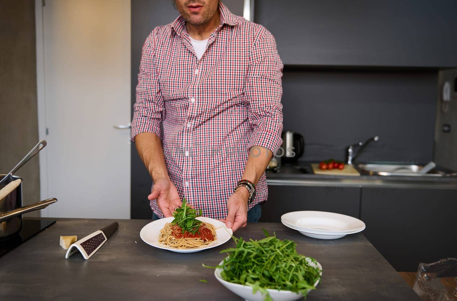 Close-up view of a young man cooking Italian pasta spaghetti for dinner in the home kitchen. Focus on served plate with Italian spaghetti with tomato sauce and greens arugula herbs