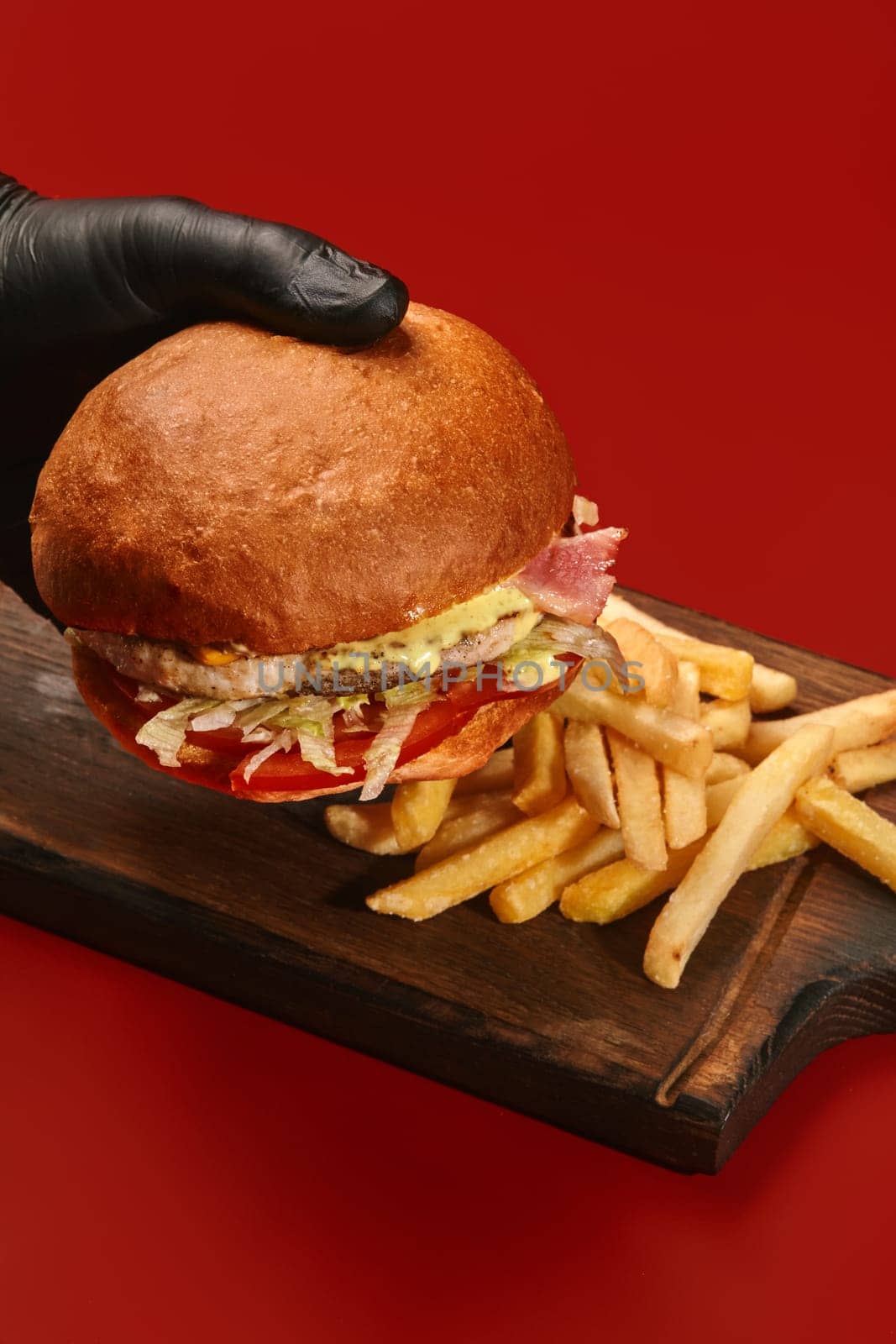 Hand in black gloves holding classic cheeseburger with juicy chicken patty, fried bacon, melted cheese and fresh vegetables served with side of golden fries on wooden board on red background