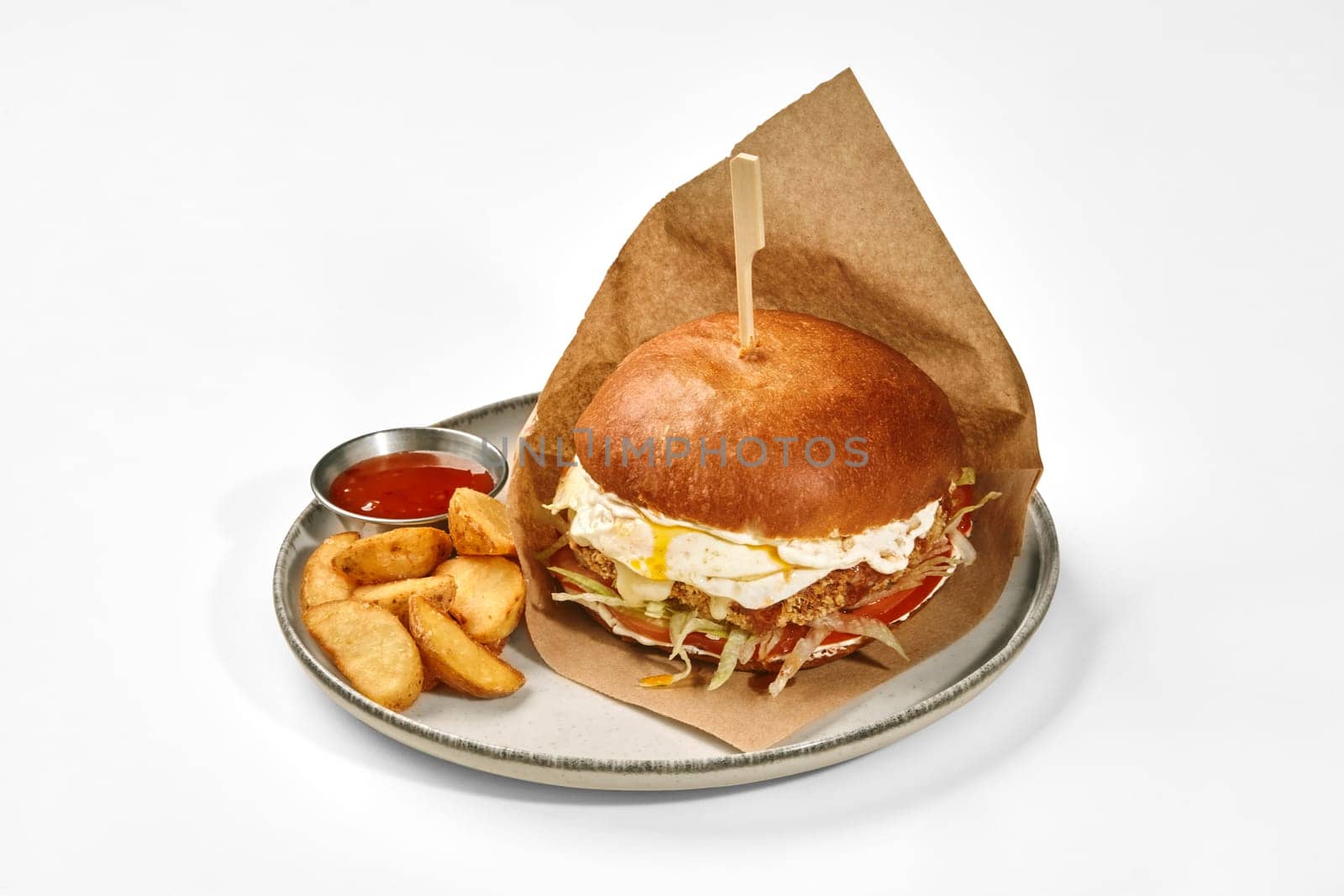 Hearty hamburger in browned bun with breaded beef patty, fried egg, tomato slices and lettuce in paper wrap served with side of baked country style potatoes and spicy sauce on ceramic plate