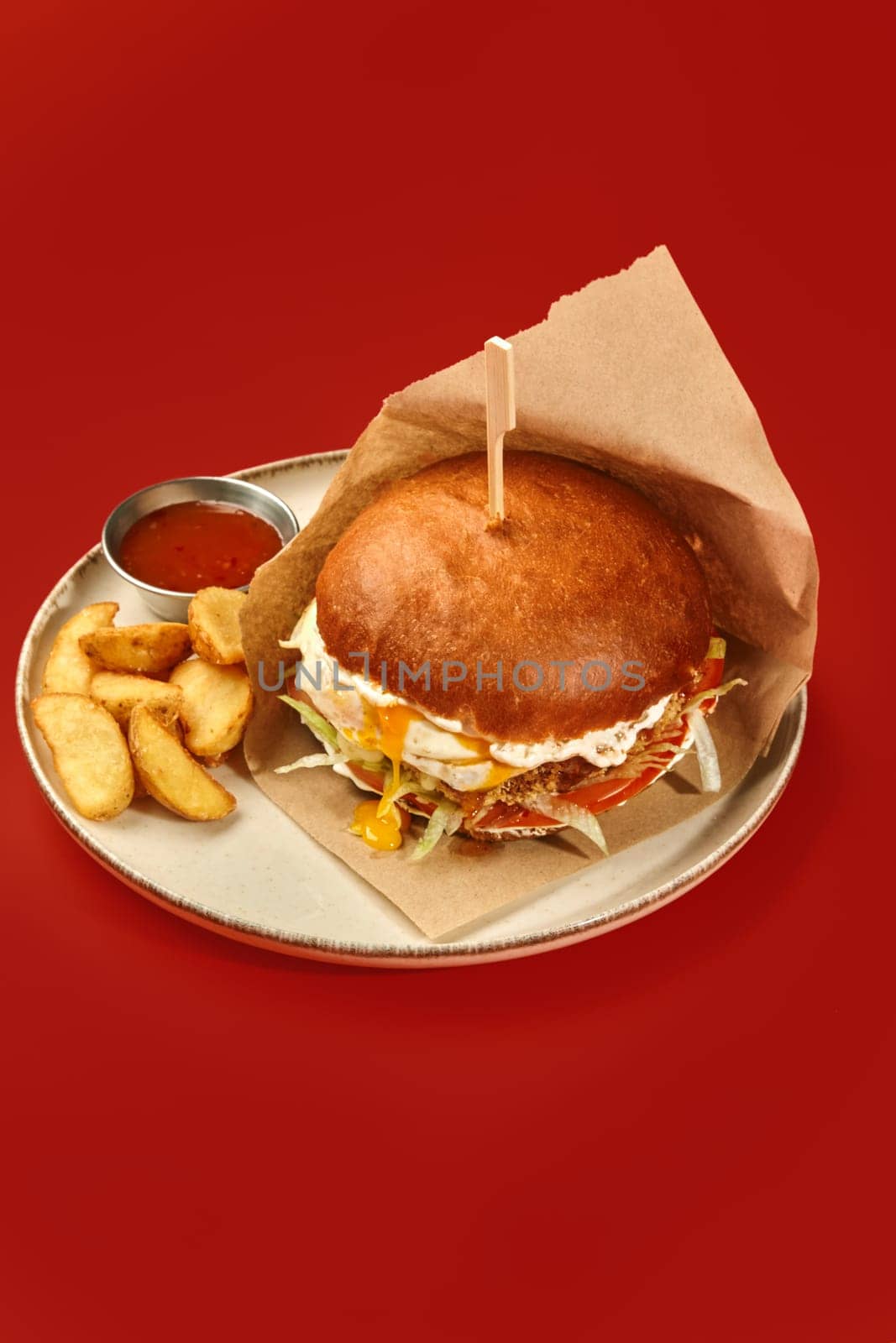 Craft-style chicken burger with fried egg and vegetables in brown paper wrap with wooden skewer served with wedge fries and dipping sauce on plate, on red background. Popular fast food concept