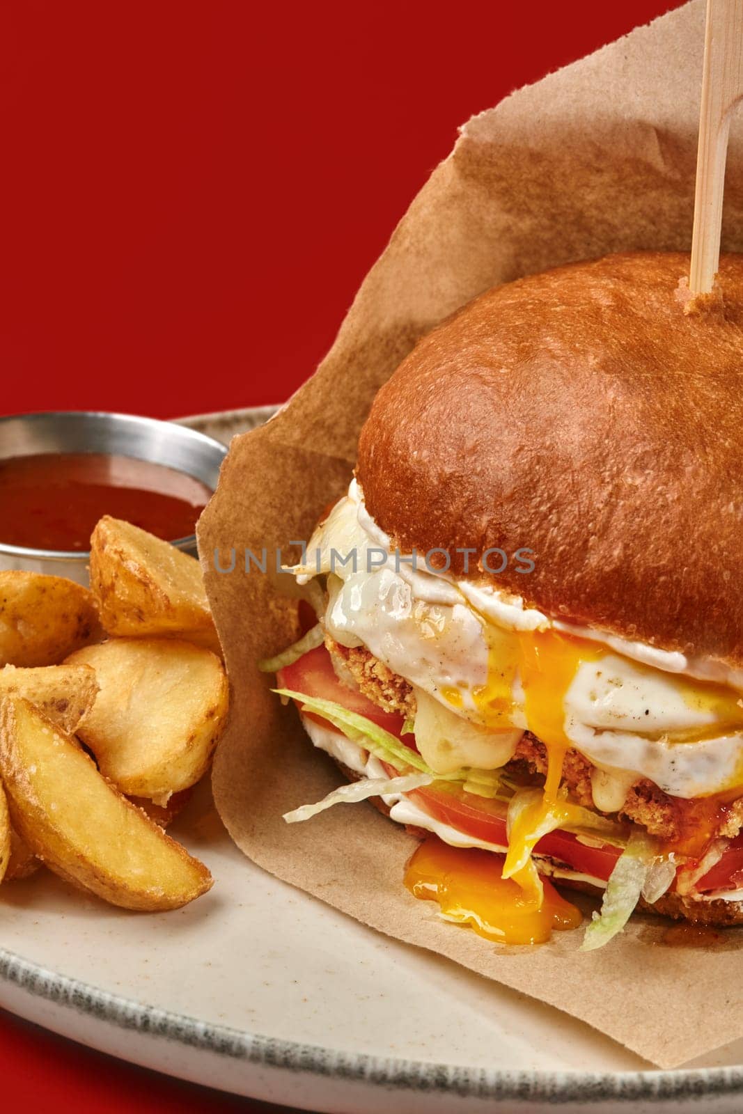Closeup of juicy beef burger in browned bun with oozing sunny side up egg, tomato slices and lettuce wrapped in craft paper, served with potato wedges and dipping sauce on plate against red backdrop