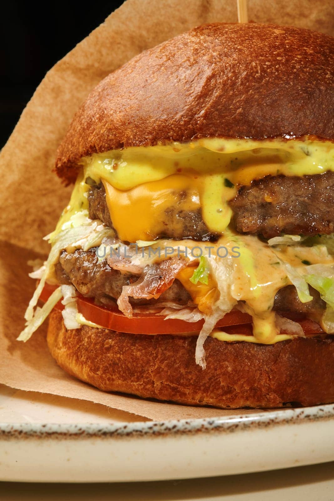 Close-up of appetizing burger in browned bun with two juicy beef patties, fried bacon slices, fresh tomatoes, lettuce and cheese sauce in craft paper wrapping on plate, against black background