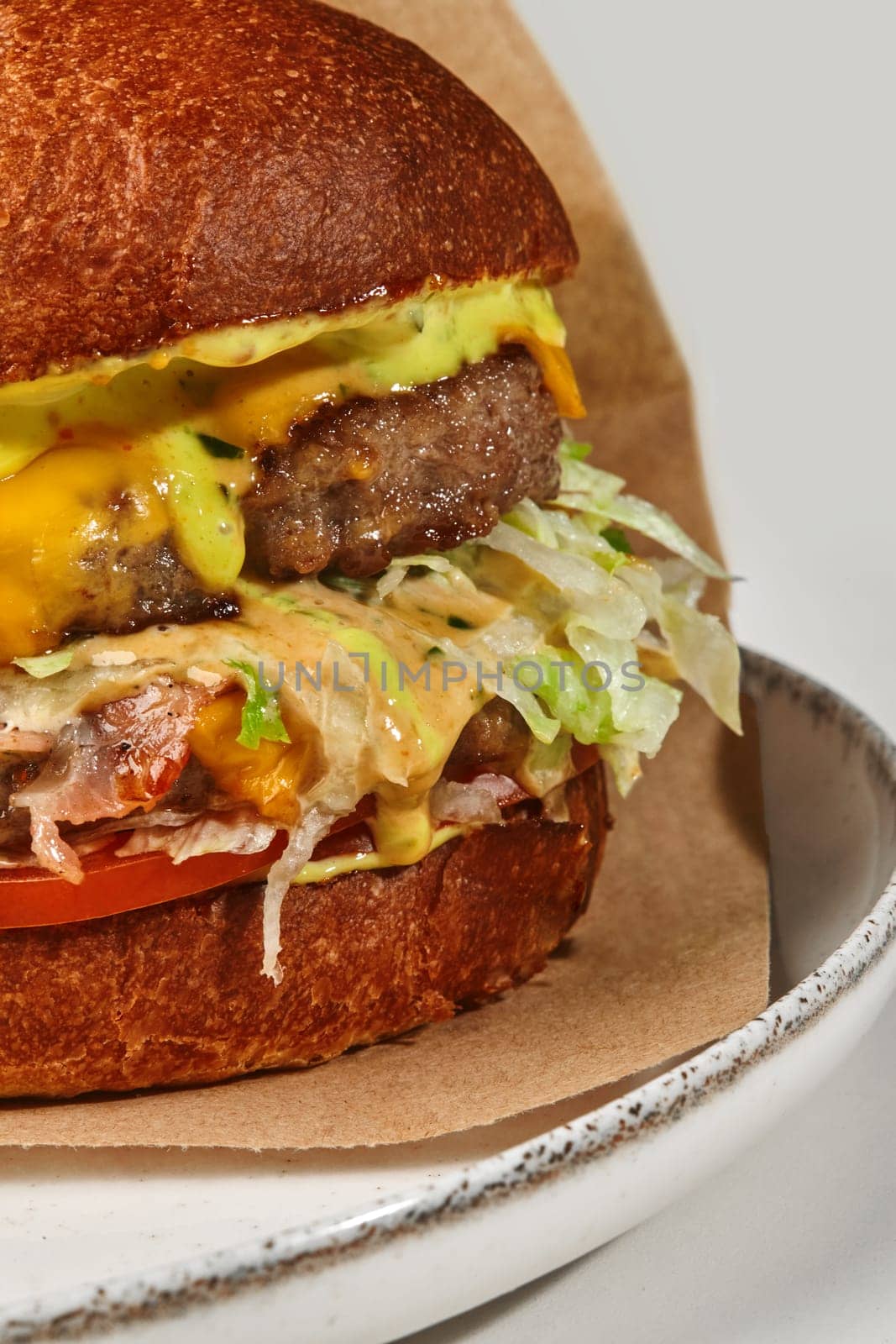 Close-up of delicious double hamburger in fluffy bun with two juicy beef patties, fried bacon slices, fresh tomatoes, lettuce and cheese sauce in craft paper wrapper on plate, against white background
