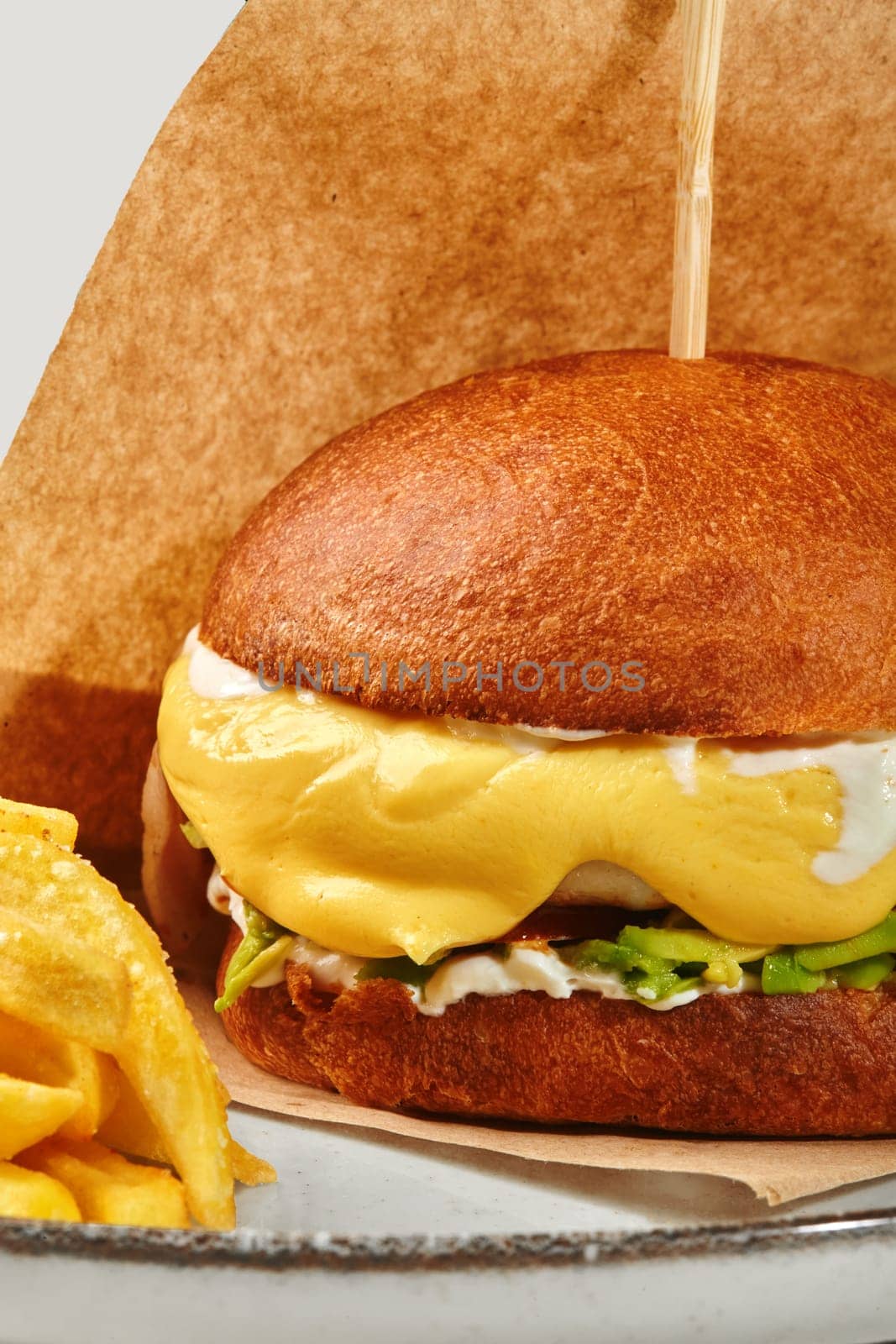 Close-up of delicious burger in browned bun with chicken patty, avocado and creamy cheese sauce in craft paper wrapper served on plate with golden fries