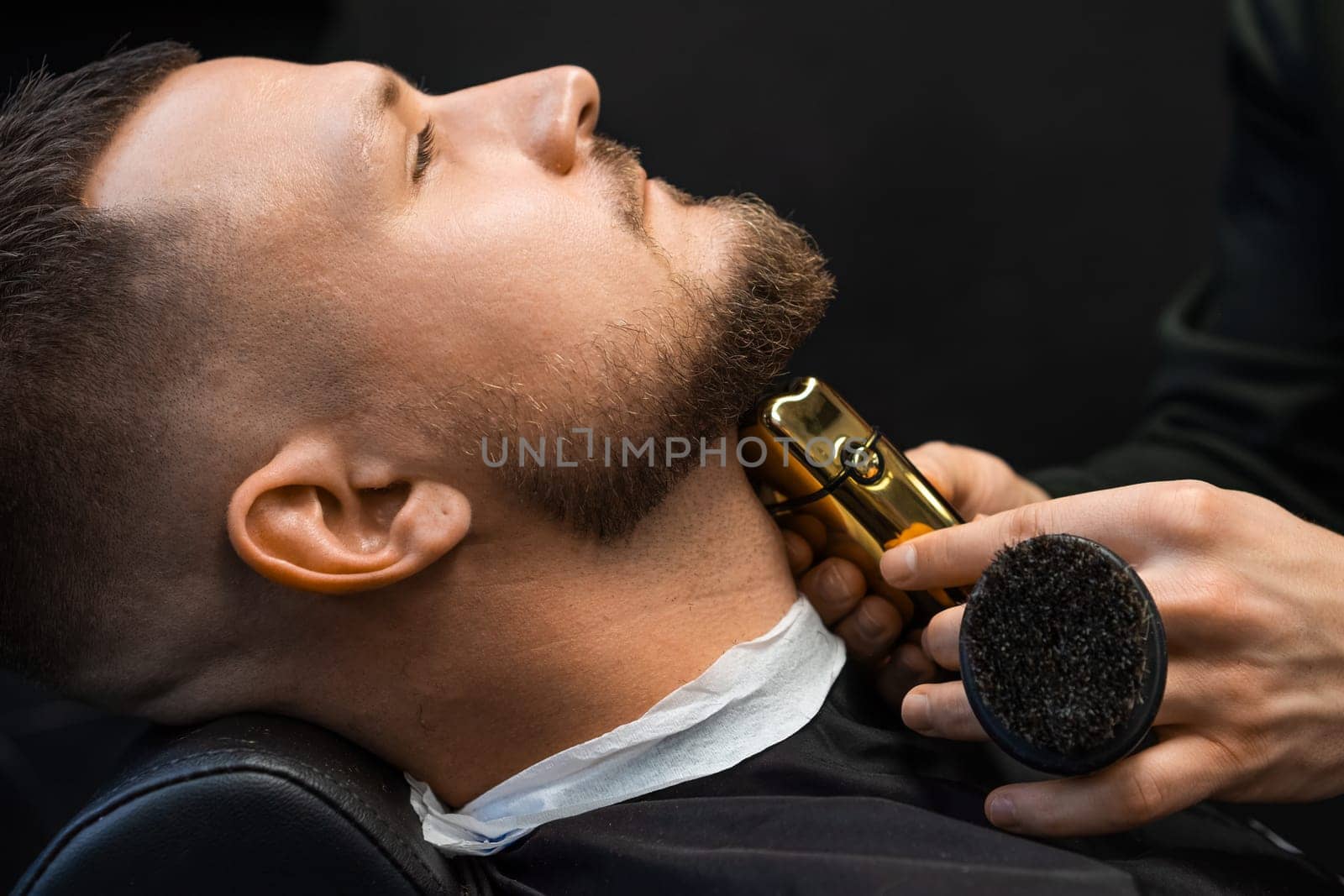 Hairdresser shaves hair on client neck creating beard shape with machine in barbershop closeup. Barber uses electrical shaver in salon