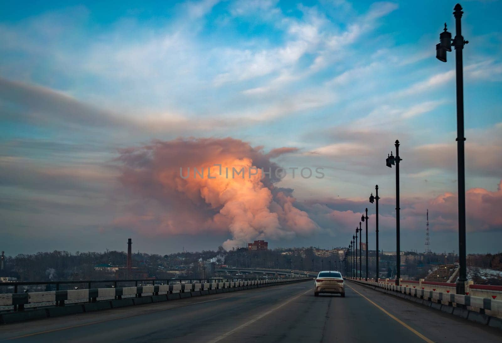 A car is driving across a bridge during sunset, with vibrant clouds and smoke rising in the distance, indicating industrial activity.