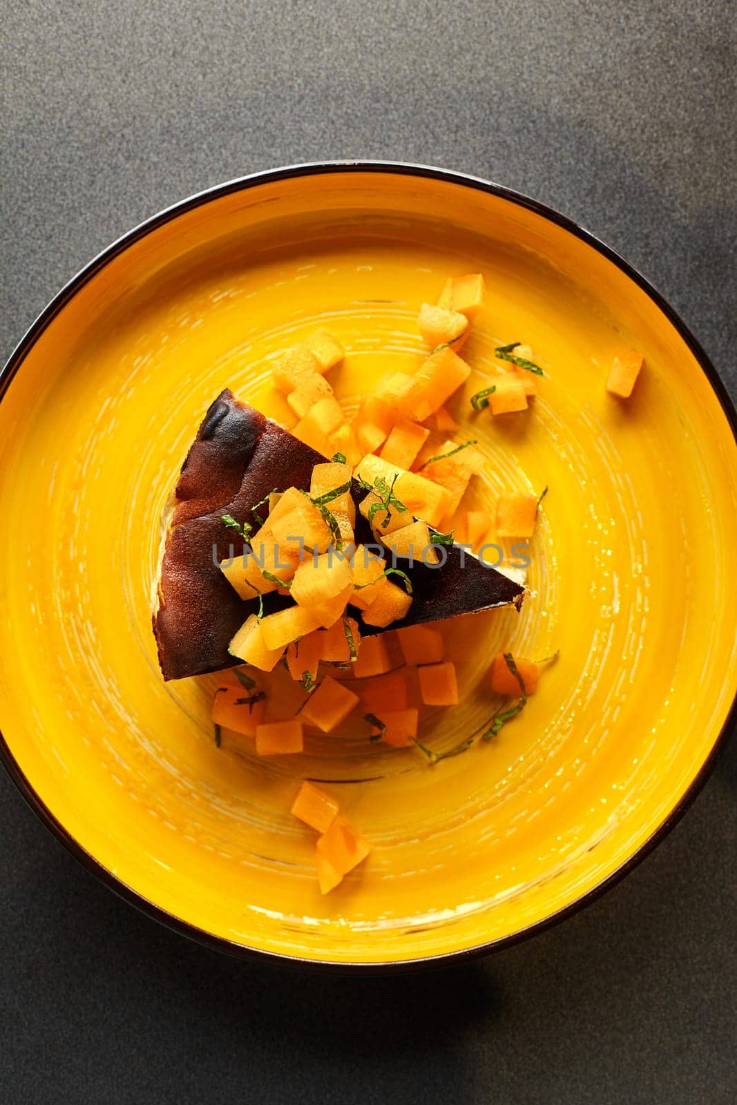 Rich caramelized burnt Basque cheesecake garnished with diced persimmon and fresh mint, displayed on bold yellow plate. Popular pastries concept