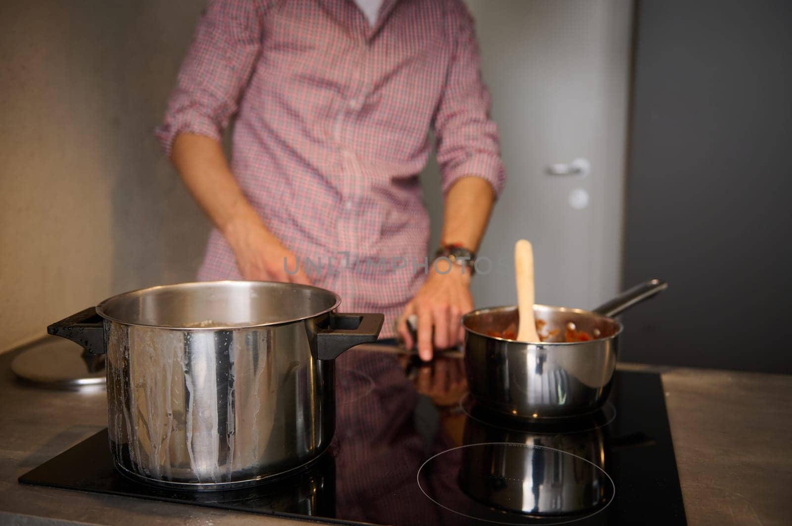 Focus on steel saucepan on the induction oven, electric stove against blurred male chef switching on panel for boiling water. Cropped view of blurred man switching on, turning on the electric stove