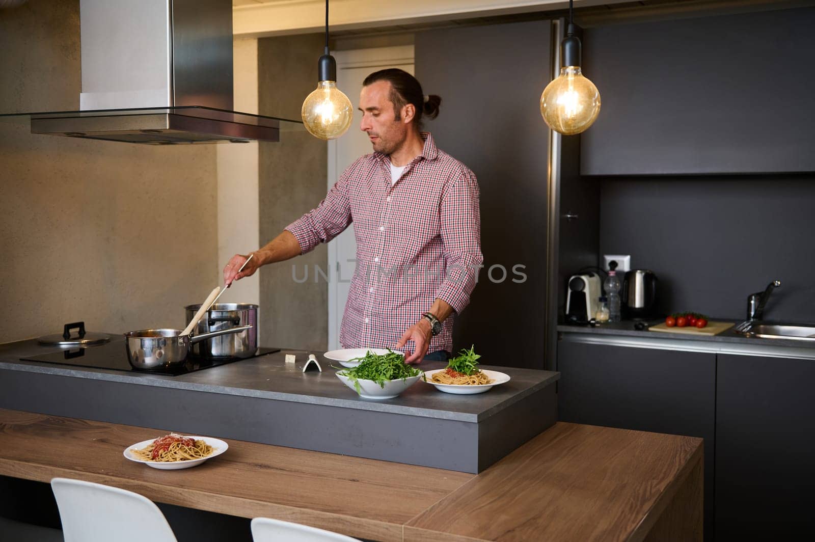 Authentic portrait of a handsome man cooking dinner at home, standing at kitchen counter, mixing ingredients in a stainless steel saucepan on the panel of induction stove. People. Culinary. Lifestyle