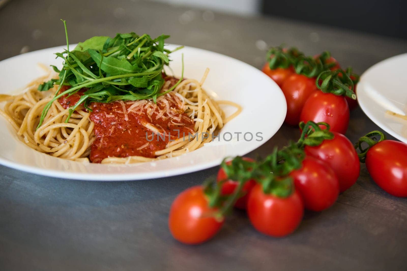 Food background. Still life with a white plate with Italian spaghetti capellini with tomato sauce, garnished with arugula green leaves and branch of red ripe organic tomatoes on the gray kitchen table