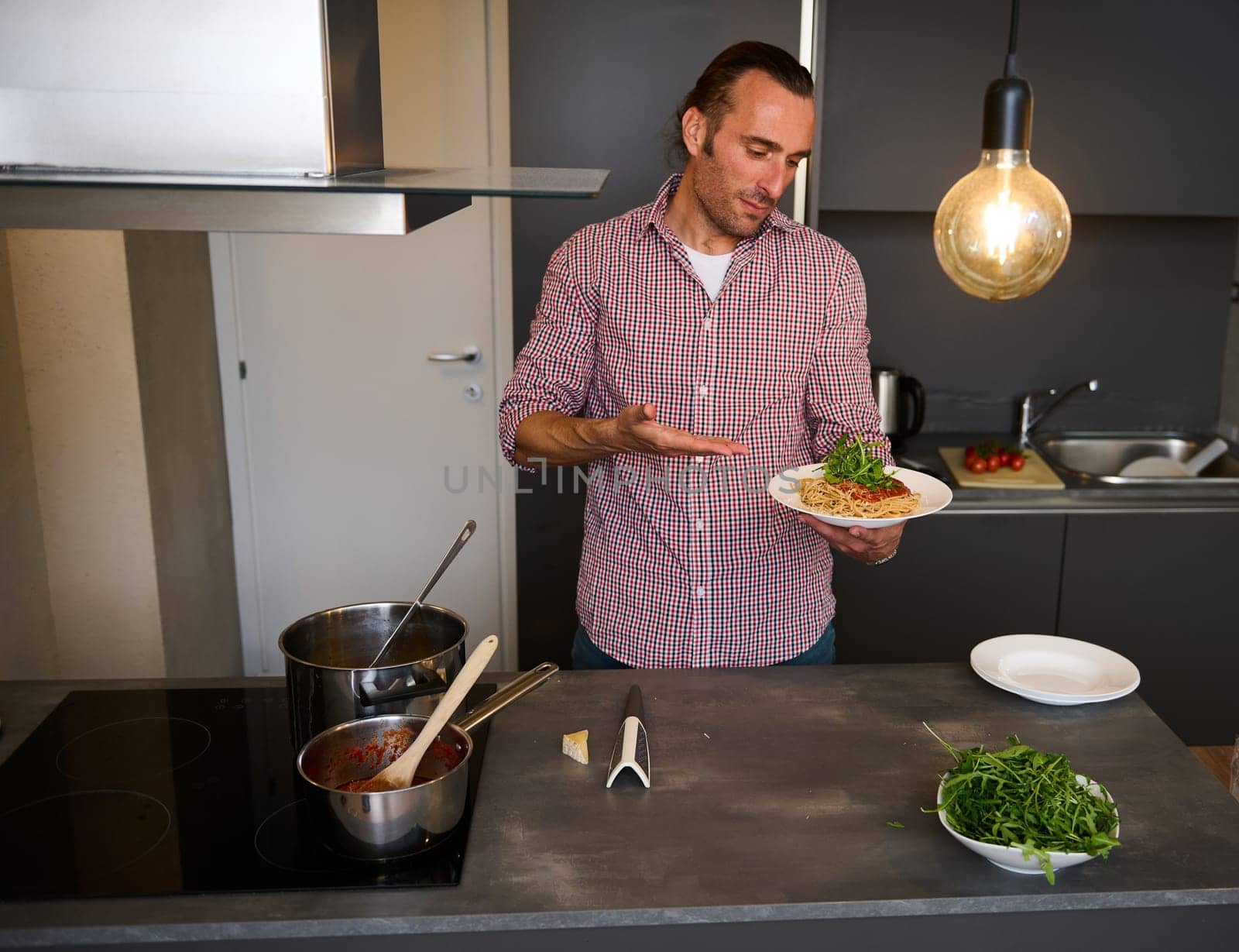 Handsome young man, Italian chef showing a white plate with Italian spaghetti capellini, standing in the home kitchen. Chef showing a served dish with Italian pasta. Italy. Food. Culture. Traditions