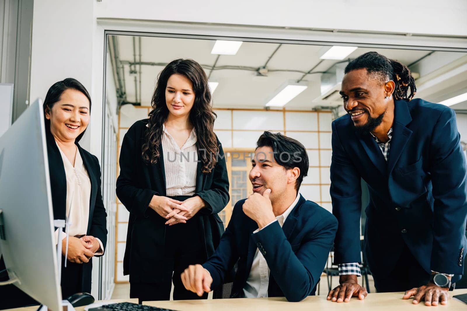 In a collaborative meeting, coworkers gather around a desktop computer, actively discussing strategy and brainstorming ideas. Their diverse backgrounds contribute to successful teamwork and planning. by Sorapop