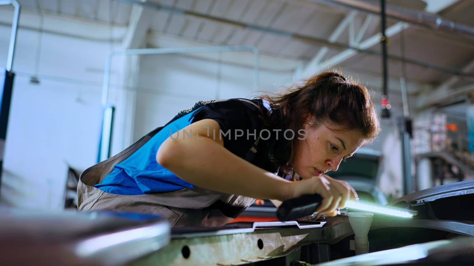 POV shot of mechanic opening car hood in auto repair shop, using work light to check for damages during routine maintenance. Garage workspace employee using professional tool to look inside vehicle