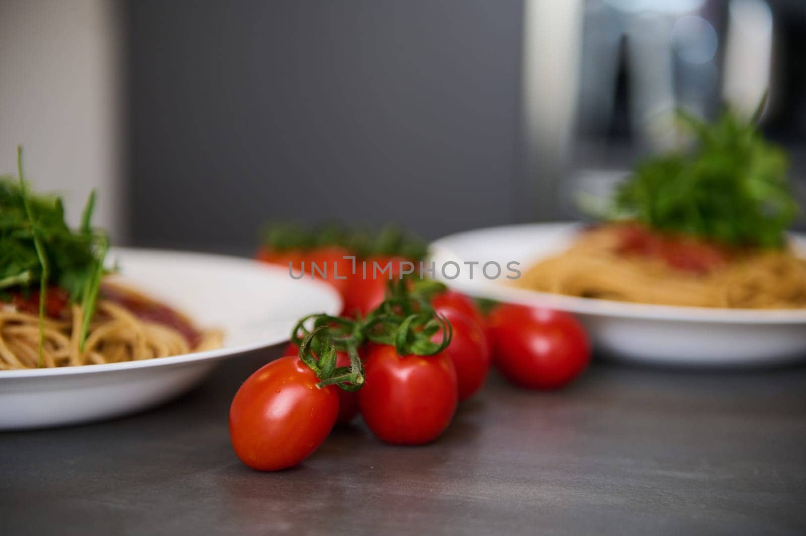 Details on yummy bunch of ripe organic cherry tomatoes, near two white plates with Italian spaghetti pasta garnished with green leaves of arugula, on kitchen table. Food background. Italian cuisine