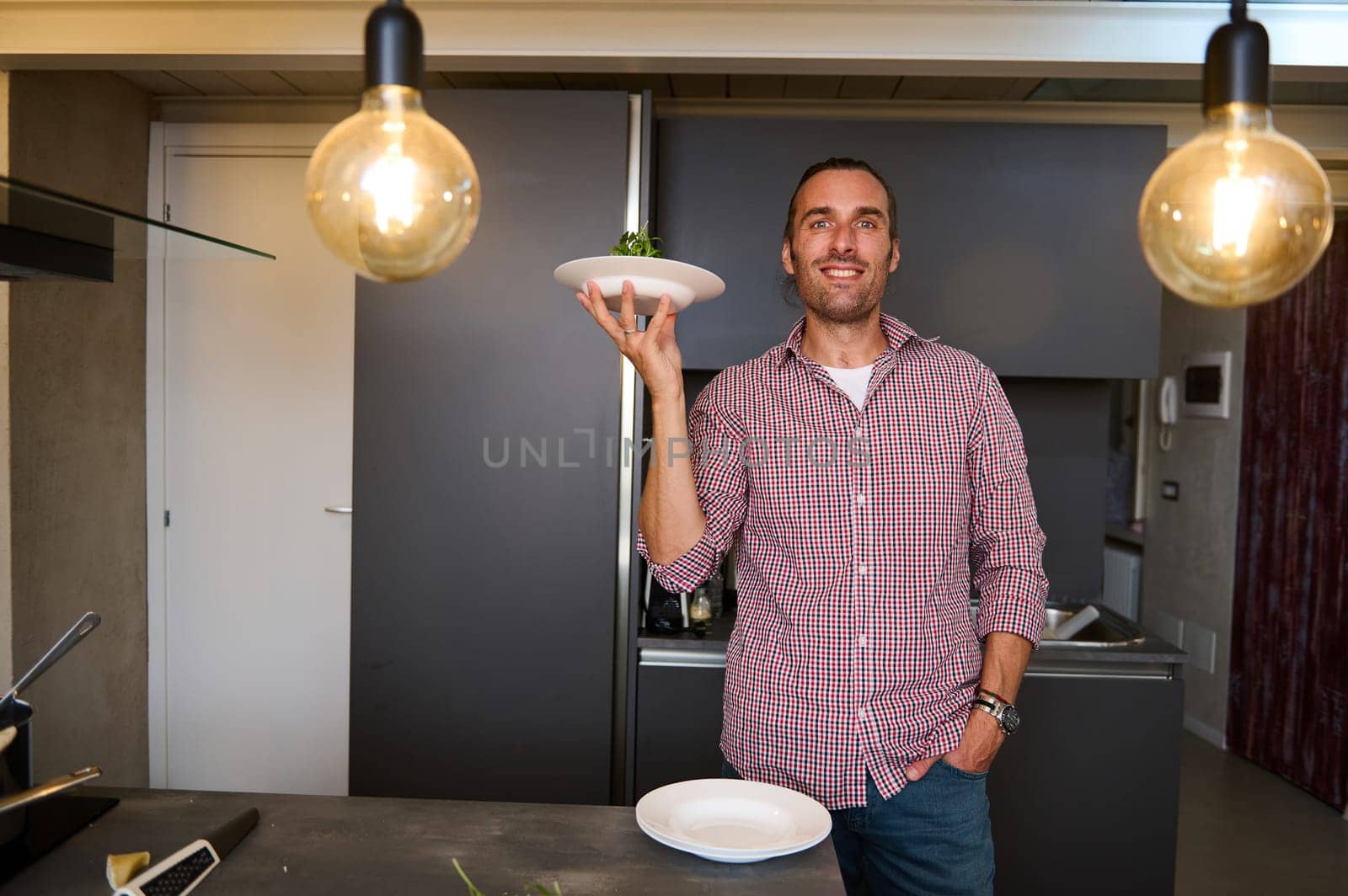 Authentic portrait of a young male chef holding a plate with Italian pasta, smiling looking at camera, standing in the minimalist home kitchen interior. People. Italian culture. Lifestyle. Culinary