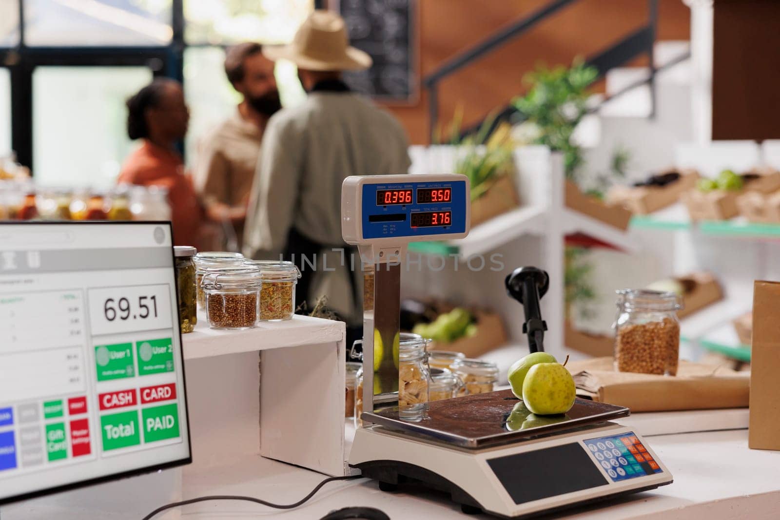 Fresh apples are placed on a digital measuring scale while multicultural customers are assisted by the salesman in background. Photo focus on cashier desk displaying weighing equipment and desktop pc.