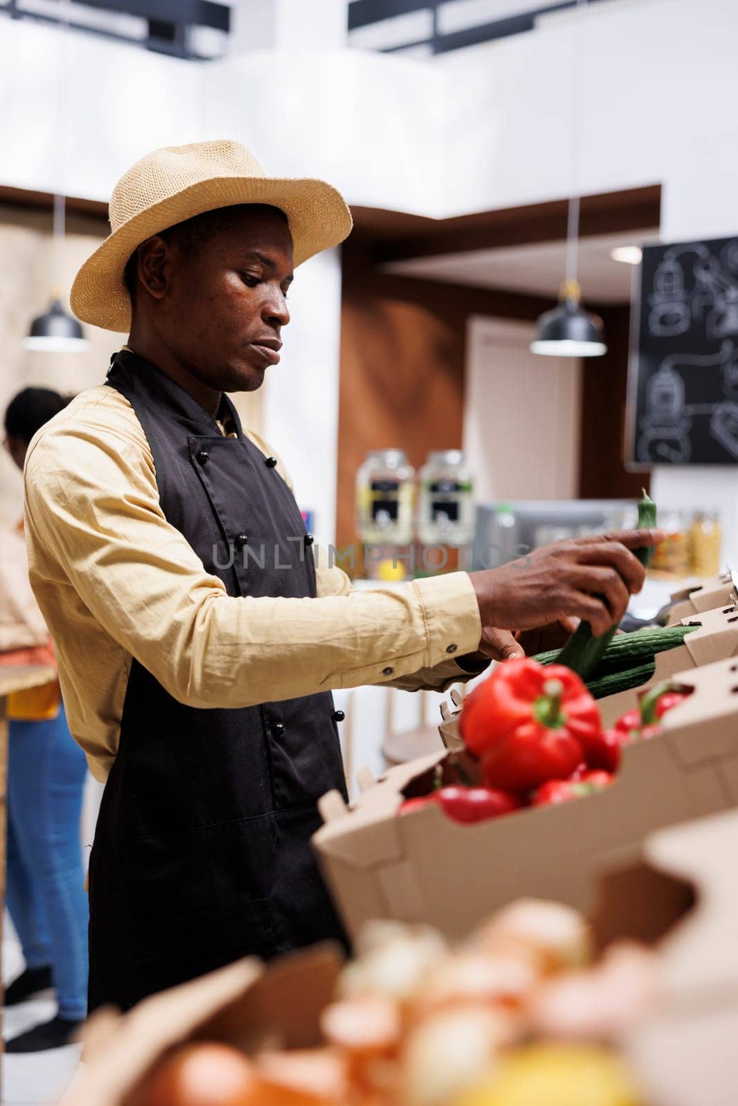 African american man with an apron organizing bio fruits and vegetables in the local supermarket. Selective focus on vibrant red peppers, cucumbers, and onions positioned on shelves.
