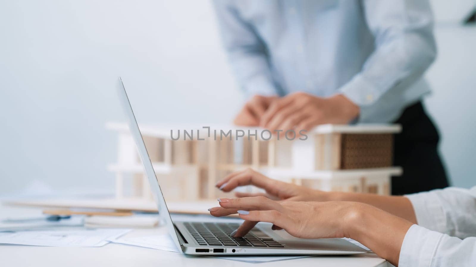 Close-up of young beautiful professional architect hands using laptop with blueprint and architectural document placed on table while male coworker inspects house model. Focus on hand. Immaculate.
