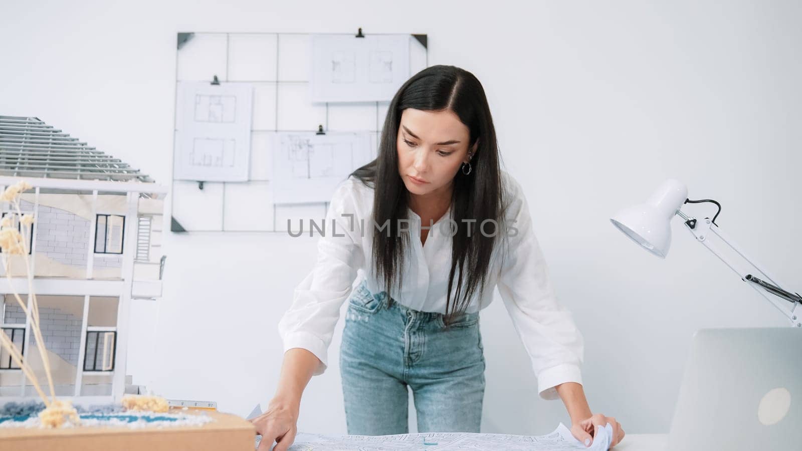 Young beautiful caucasian architect puts blueprint on the table and compare with house model while inspect hos model construction carefully at modern office with blueprint striped behind. Immaculate.