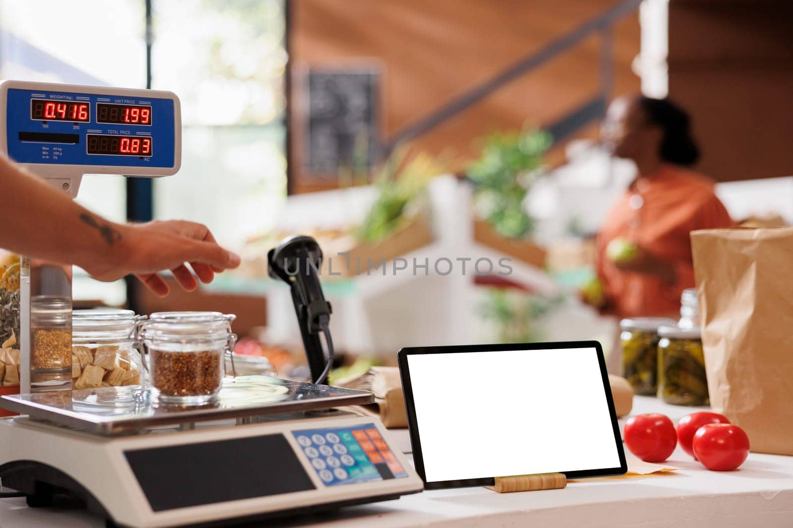 Glass jar filled with bio food items is placed on a measuring weight scale on the counter of grocery store. Image shows a tablet displaying blank chromakey mockup template while seller weighs an item.
