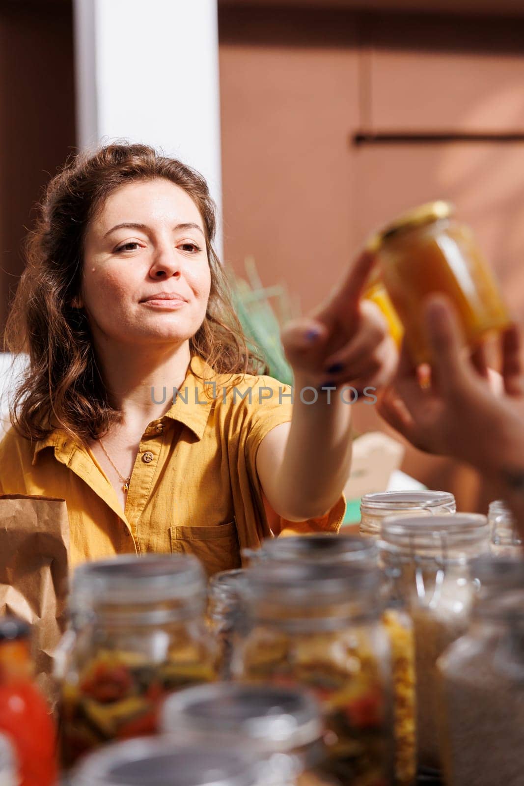 Storekeeper showing woman shopping in zero waste grocery store organic nutritious honey in reusable packaging. Vendor in ethically conscious shop recommending food item to customer