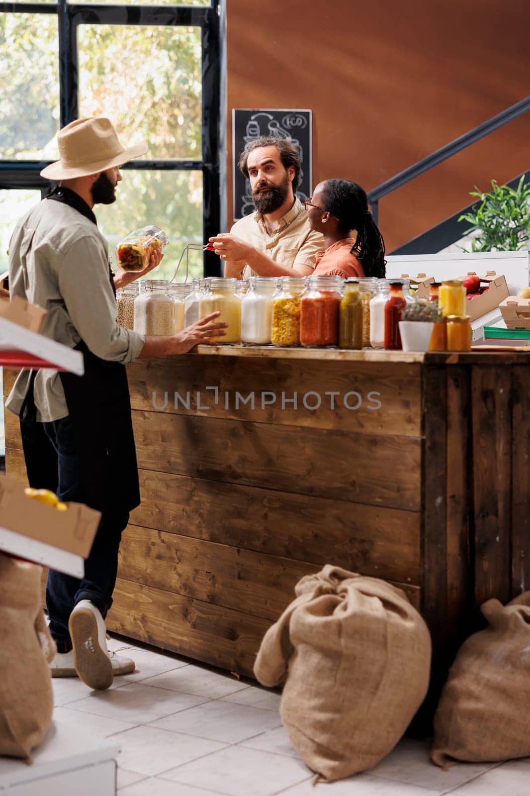 Caucasian man and black woman shopping for pantry staples in sustainable zero waste store, being assisted with advice from male vendor. Local supermarket owner showing couple bio items.