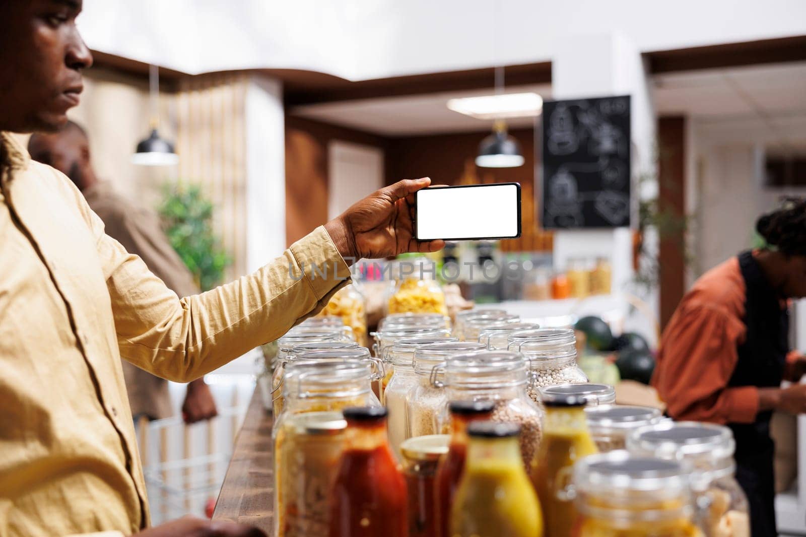 At eco friendly grocery shop African American male consumer holds a cell phone that displays an isolated white screen. Image shows black man with mobile device having a chromakey template.