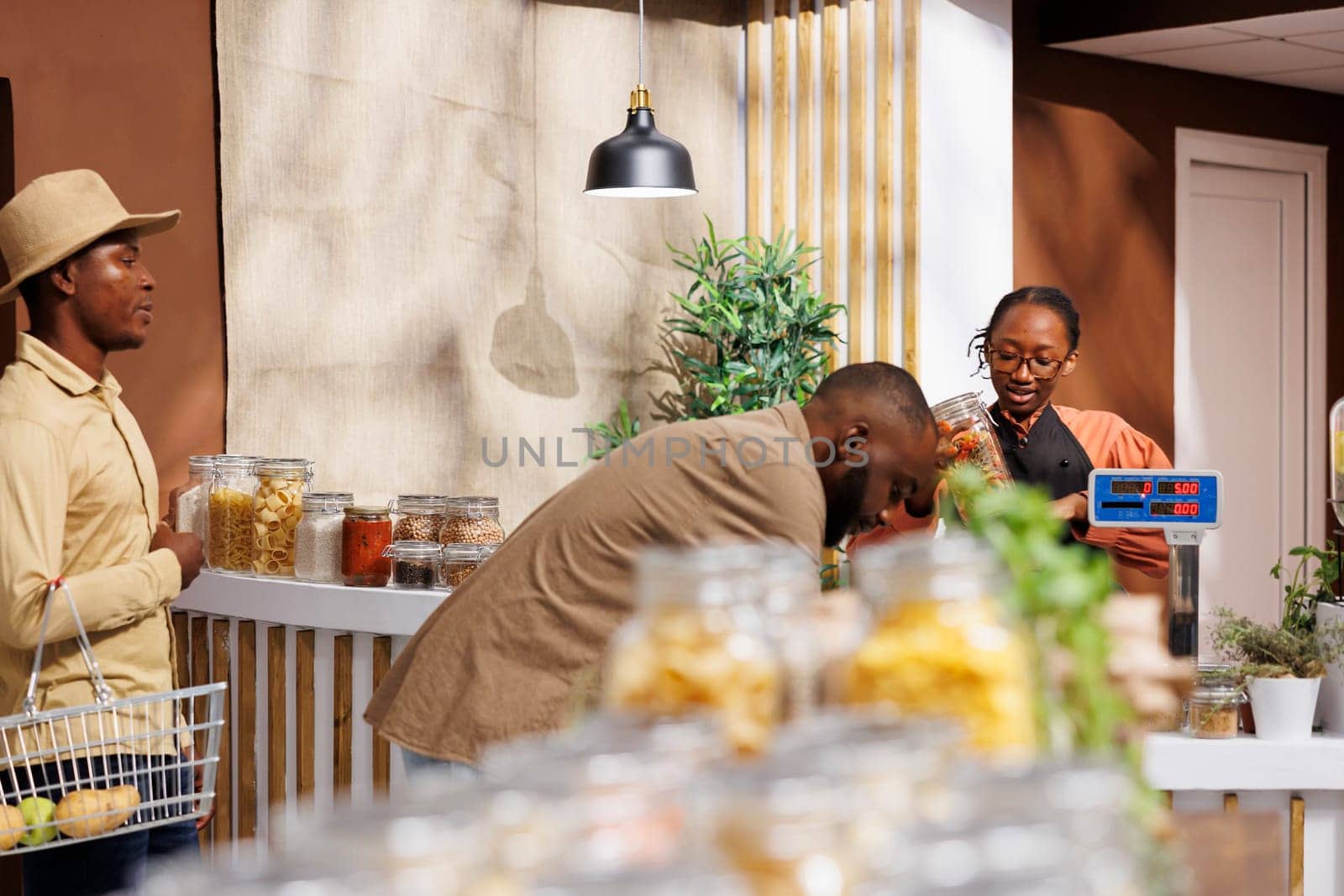 Black man wearing hat waits in line while female cashier assists with packaging of products of another male customer. Image shows African American clients at checkout counter of eco friendly store.