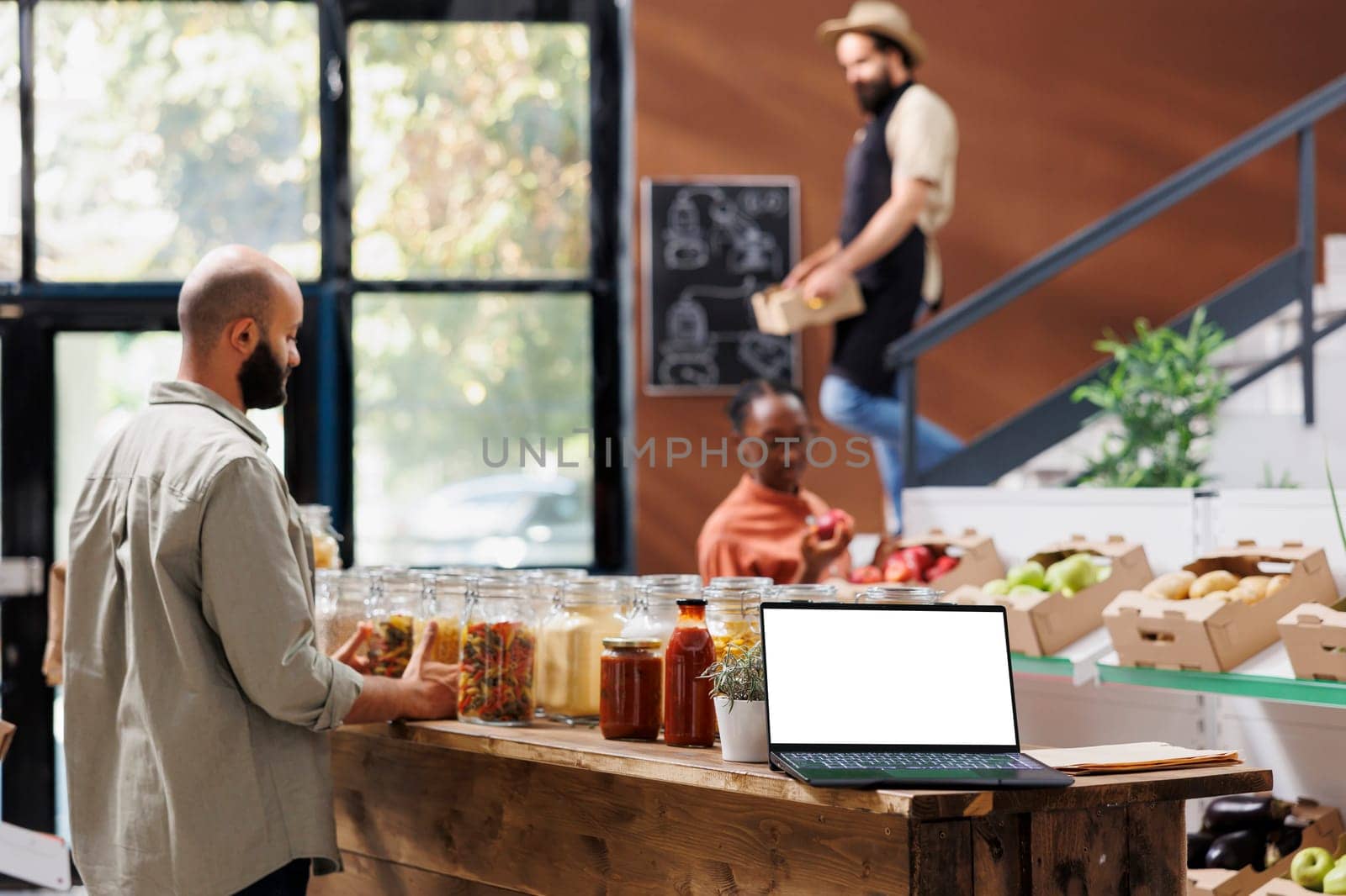 Wireless computer with isolated chromakey mockup template is placed on wooden counter while customers are browsing in the store. Laptop is displaying a blank white screen for advertisements.