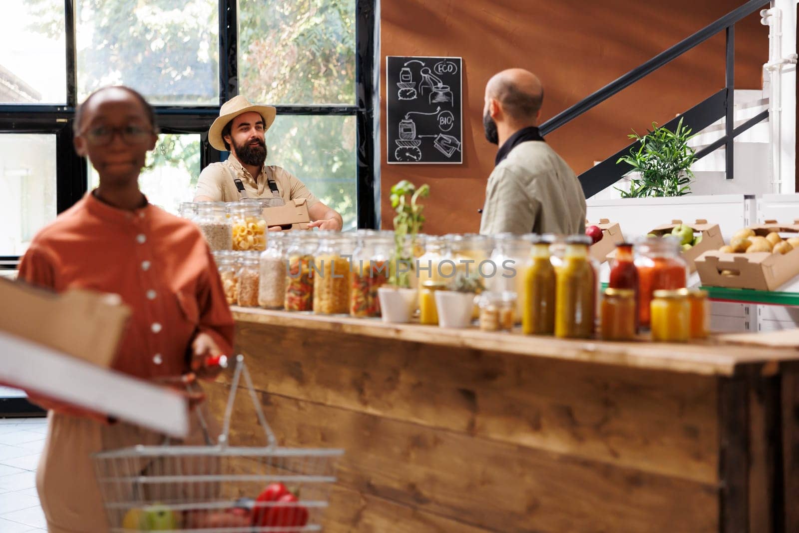 Black woman looks around in store while holding basket filled with food products. Focus on background are two men, one a farmer with boxes and another a store worker, interacting with each other.