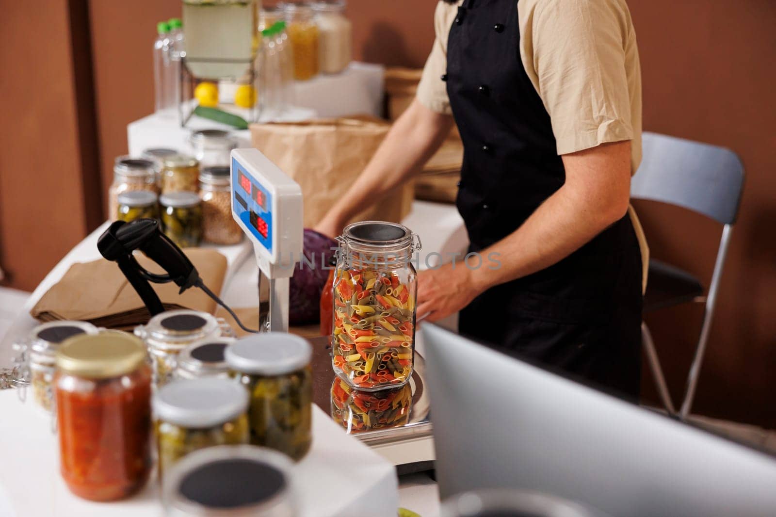 Storekeeper weighs and measures glass jar filled with colorful bio food products, on a weight scale. Male cashier wears a black apron and monitors the values showing up on digital measuring equipment.