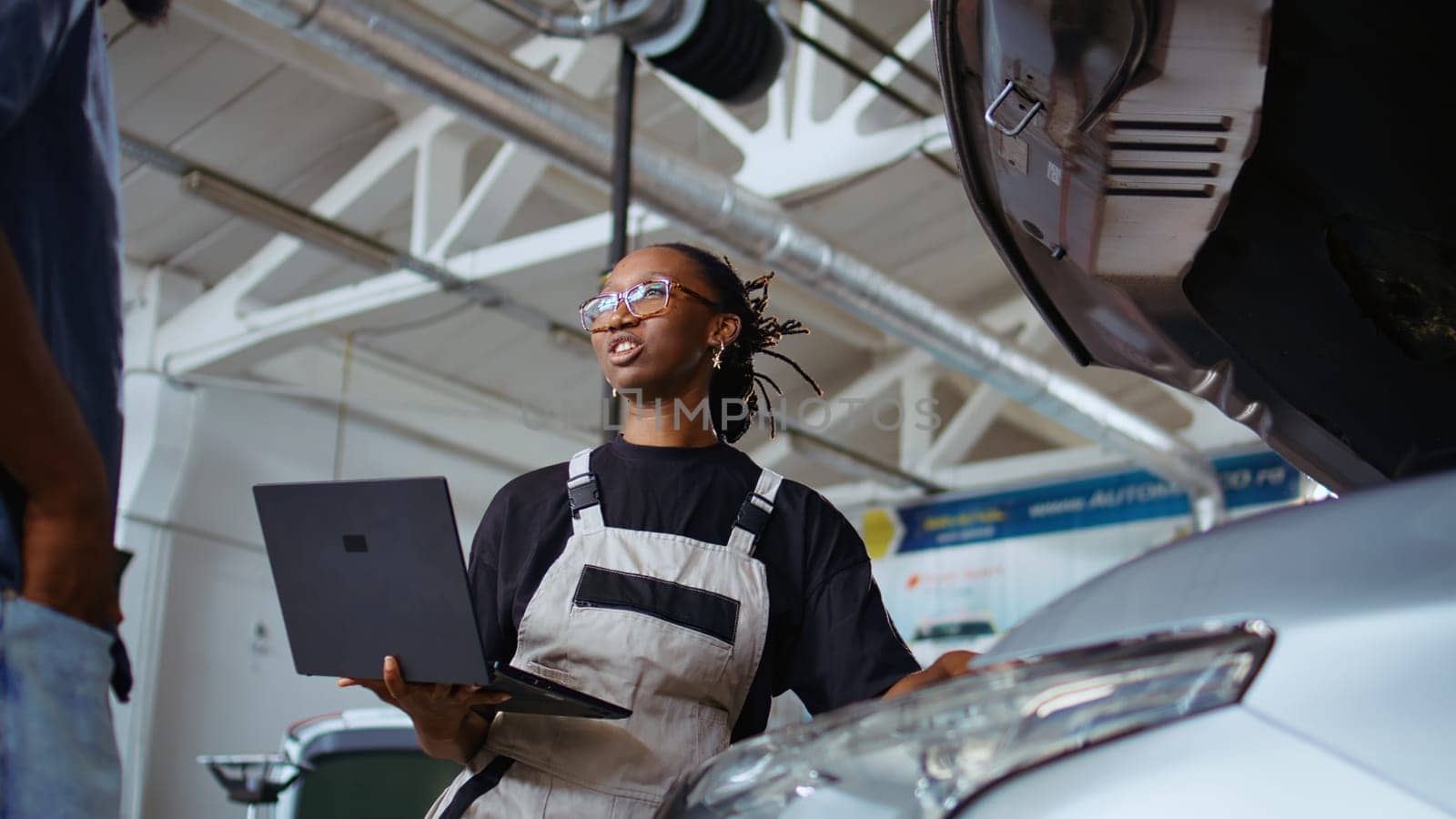 Experienced technician listens to customer in repair shop, using laptop to write down car modifications needed. Professional in garage with client, listening to her requests, zoom out shot