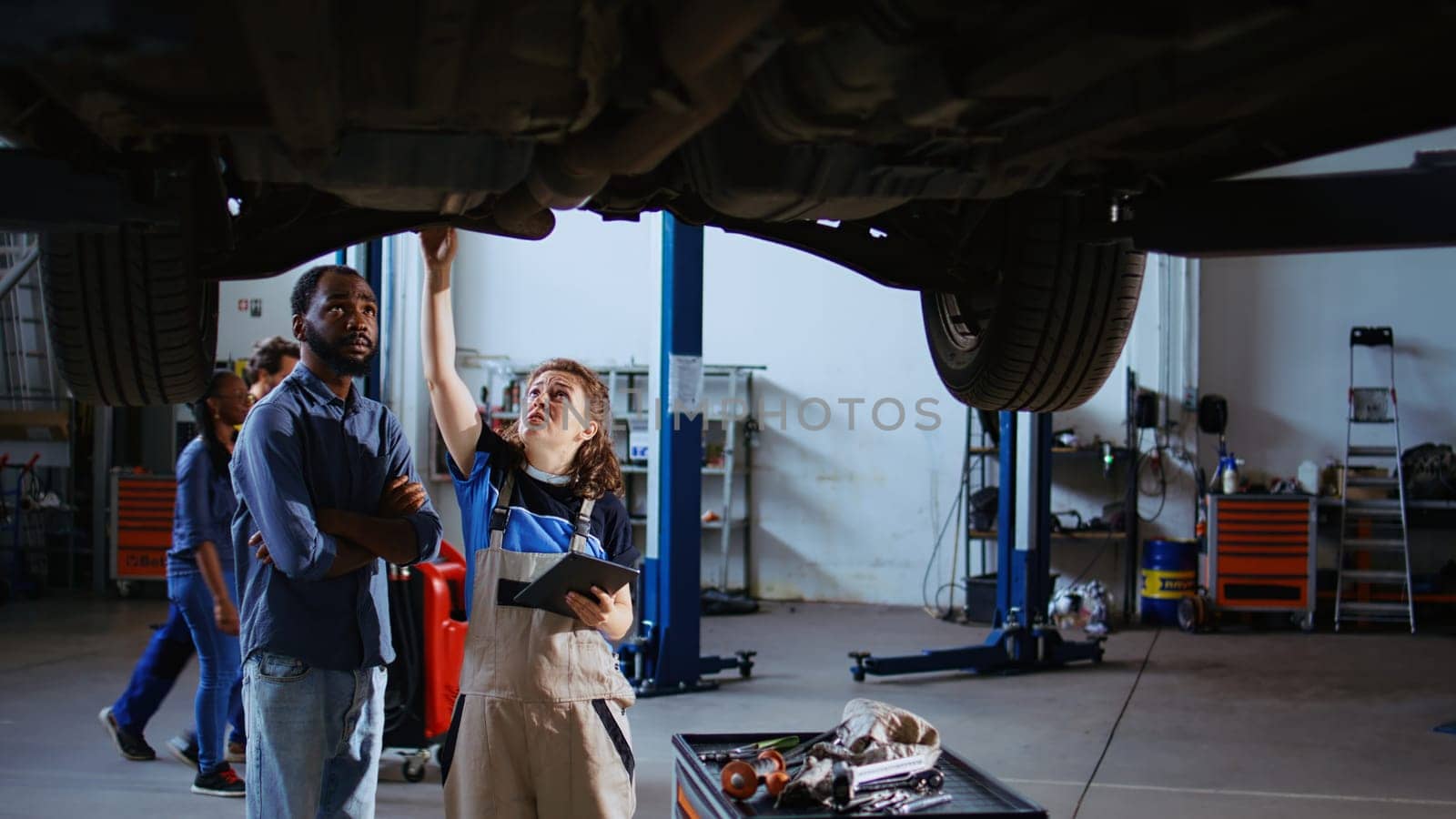 Licensed technician in repair shop showing customer what needs to be changed on his car. Garage worker inspecting vehicle placed on overhead lift, talking with BIPOC client