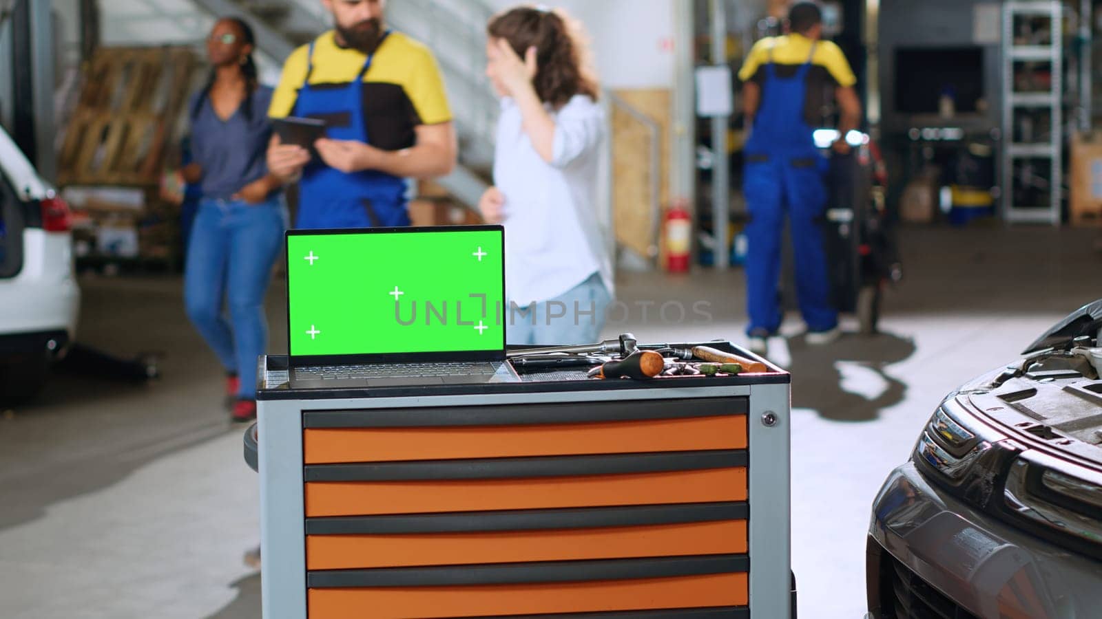 Green screen laptop placed on working bench in busy garage next to professional tools while employees roam around in blurry background. Chroma key device in auto repair shop
