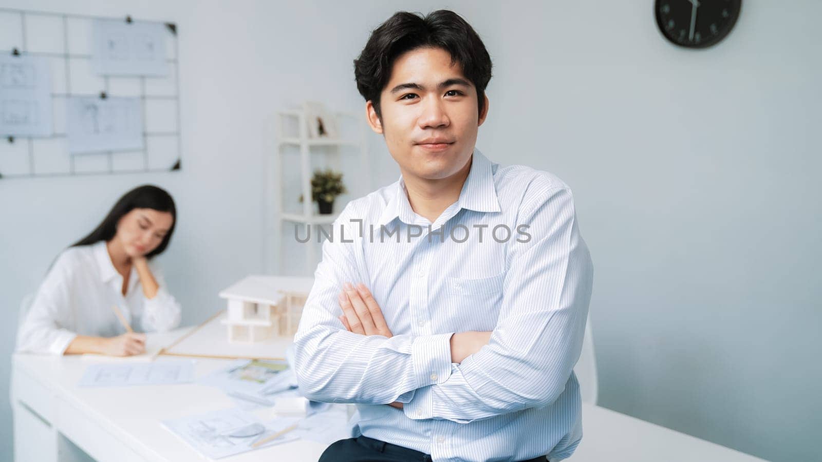 Professional asian businessman crosses arms with confident while young beautiful caucasian colleague drawing a blueprint on meeting table with house model and blueprint at modern office. Immaculate.