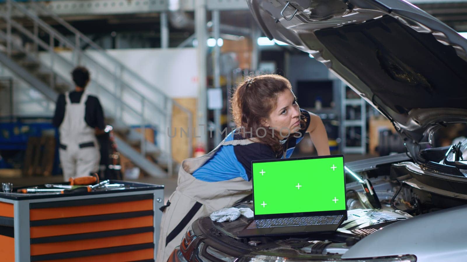 Chroma key laptop in garage facility sitting on malfunctioning car. Green screen device in auto repair shop next to mechanic cleaning vehicle inside engine compartment after fixing it