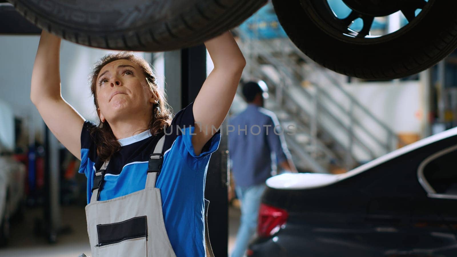 Close up shot of mechanic working on suspended car in garage, changing tires during routine maintenance. Woman in auto repair shop underneath vehicle looking to fix damages