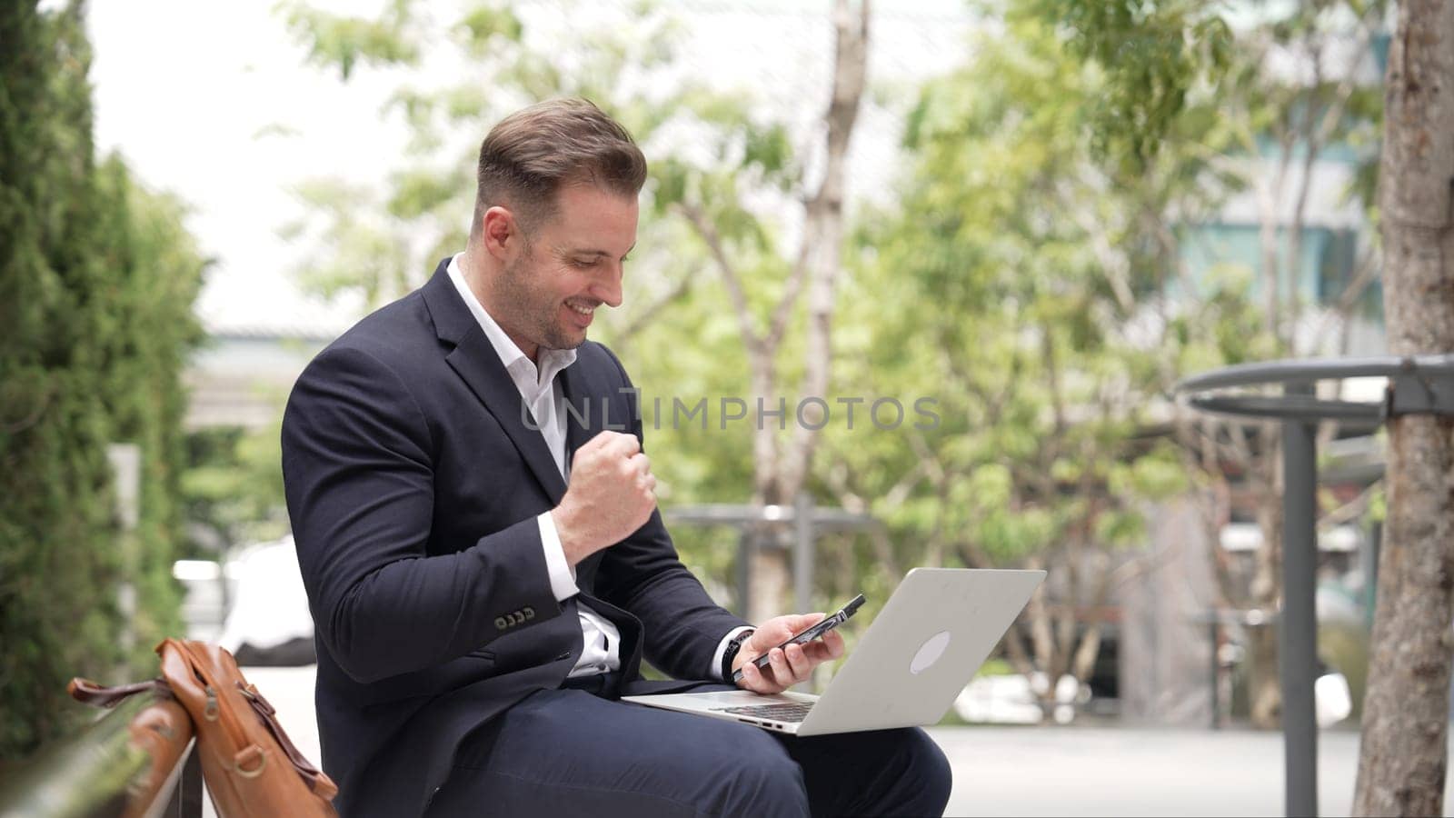Caucasian business people calling phone while sitting at green city. Happy project manager celebrate successful project or receive good news from telephone while working on laptop at park. Urbane.