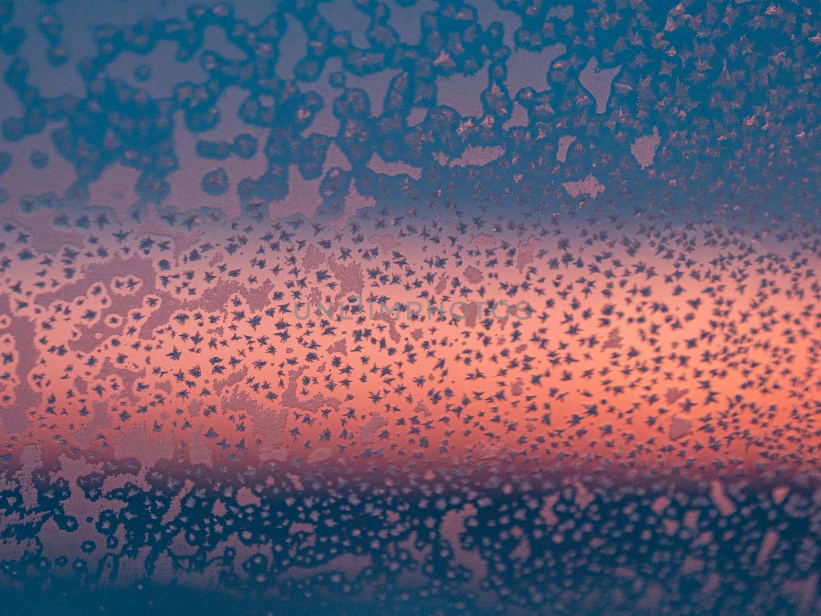 Frozen window with frost patterns overlooking colorful sunset in winter evening