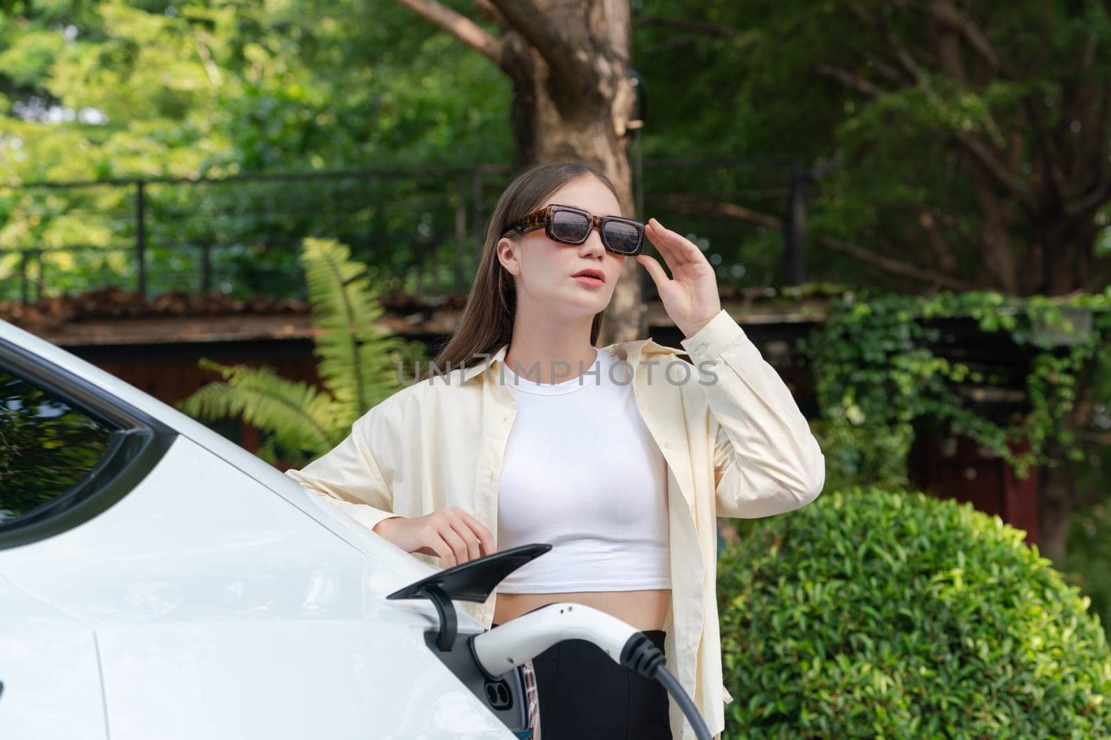 Young woman recharge her EV electric vehicle at green city park parking lot. Urban sustainability lifestyle for environmental friendly EV car with battery charging station. Expedient