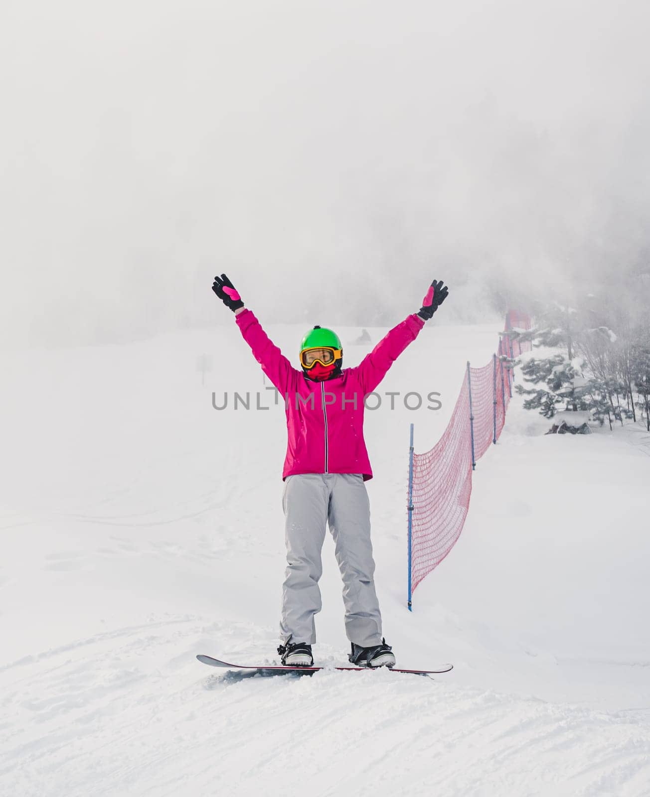 A snowboarder celebrates with arms raised on a misty, snow-covered slope. The scene is set in a winter landscape with some trees and a safety net in the background. by Busker