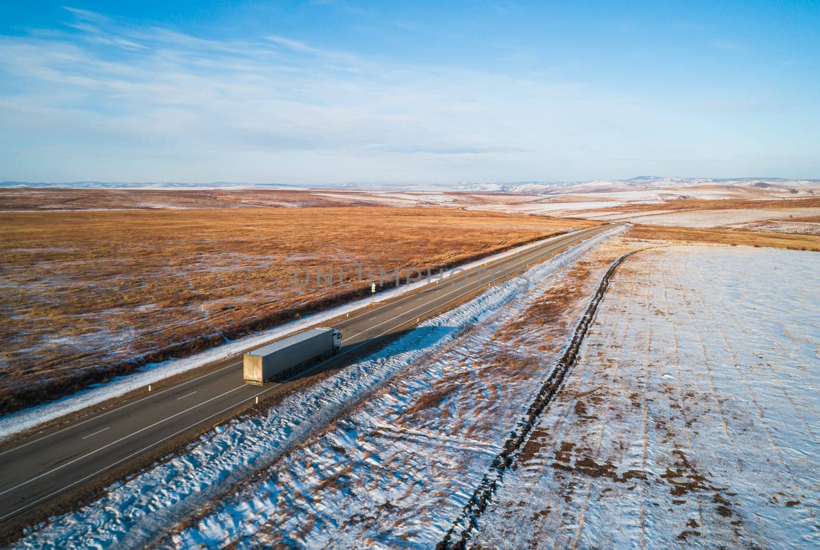 A lone semi truck drives along a winding rural highway through a snow-covered landscape during dusk in winter.